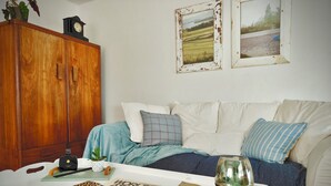 Un salon chaleureux avec un meuble en bois, un canapé blanc agrémenté de coussins à motifs, une table basse sur laquelle reposent un livre et des objets décoratifs, et des tableaux représentant des paysages, accrochés au mur.