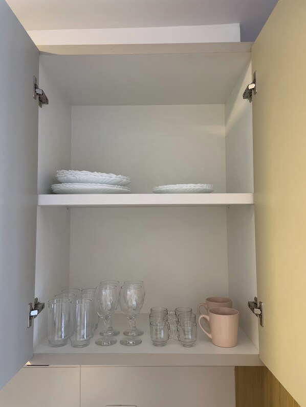 A white cabinet with two shelves; the top shelf holds stacked plates, and the bottom shelf contains various glassware and a pink mug.