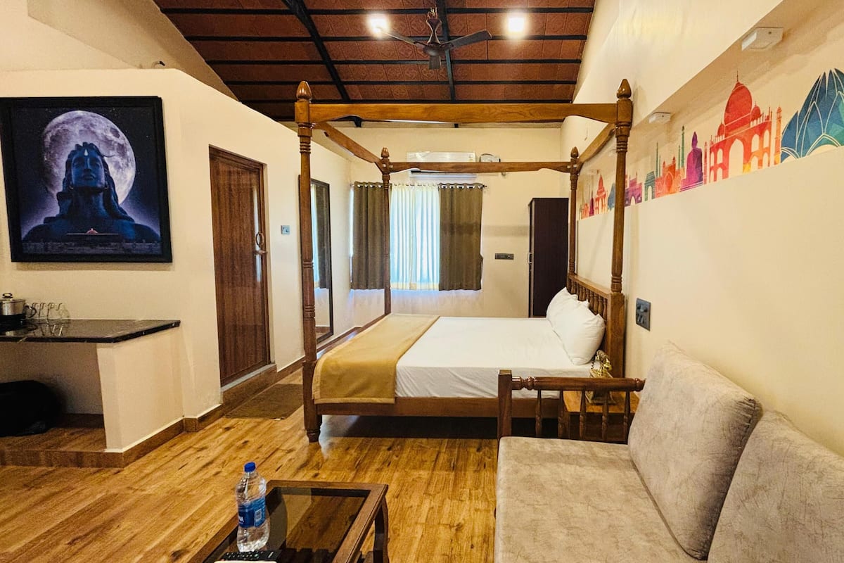 A four-poster bed with a wooden frame, a sofa, a small table with a bottle, and a wall with a colorful mural.