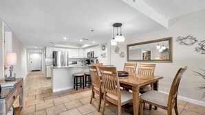 A spacious kitchen with white cabinets, a central island, and a dining area with wooden chairs and a table. - Beachfront End Unit 3-Bedroom with Wrap-Around Balcony (Indian Shores)