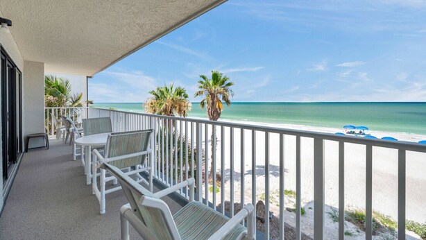A balcony with white chairs and a table, overlooking a beach with palm trees and a clear sky. - Beachfront End Unit 3-Bedroom with Wrap-Around Balcony (Indian Shores)