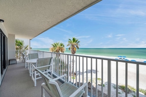 A balcony with white chairs and a table, overlooking a beach with palm trees and a clear sky. - Beachfront End Unit 3-Bedroom with Wrap-Around Balcony (Indian Shores)
