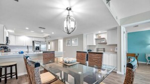 Una cocina moderna con armarios blancos, una mesa de comedor de vidrio, sillas de mimbre y una lámpara colgante.