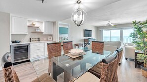 Una cocina moderna con armarios blancos, un comedor con mesa de vidrio y sillas de mimbre, y una sala de estar con sofá y televisor.
