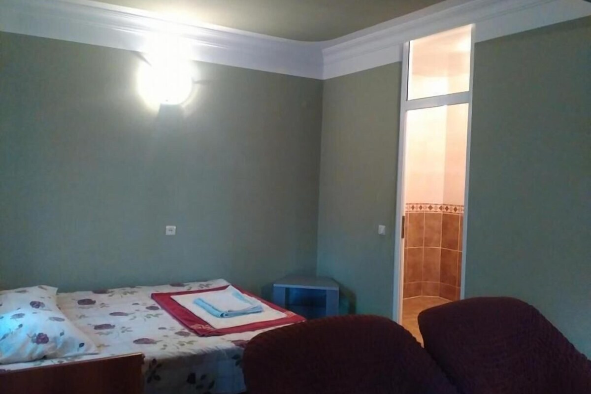 A small room with a bed, a chair, and a nightstand. There is a door leading to another room.