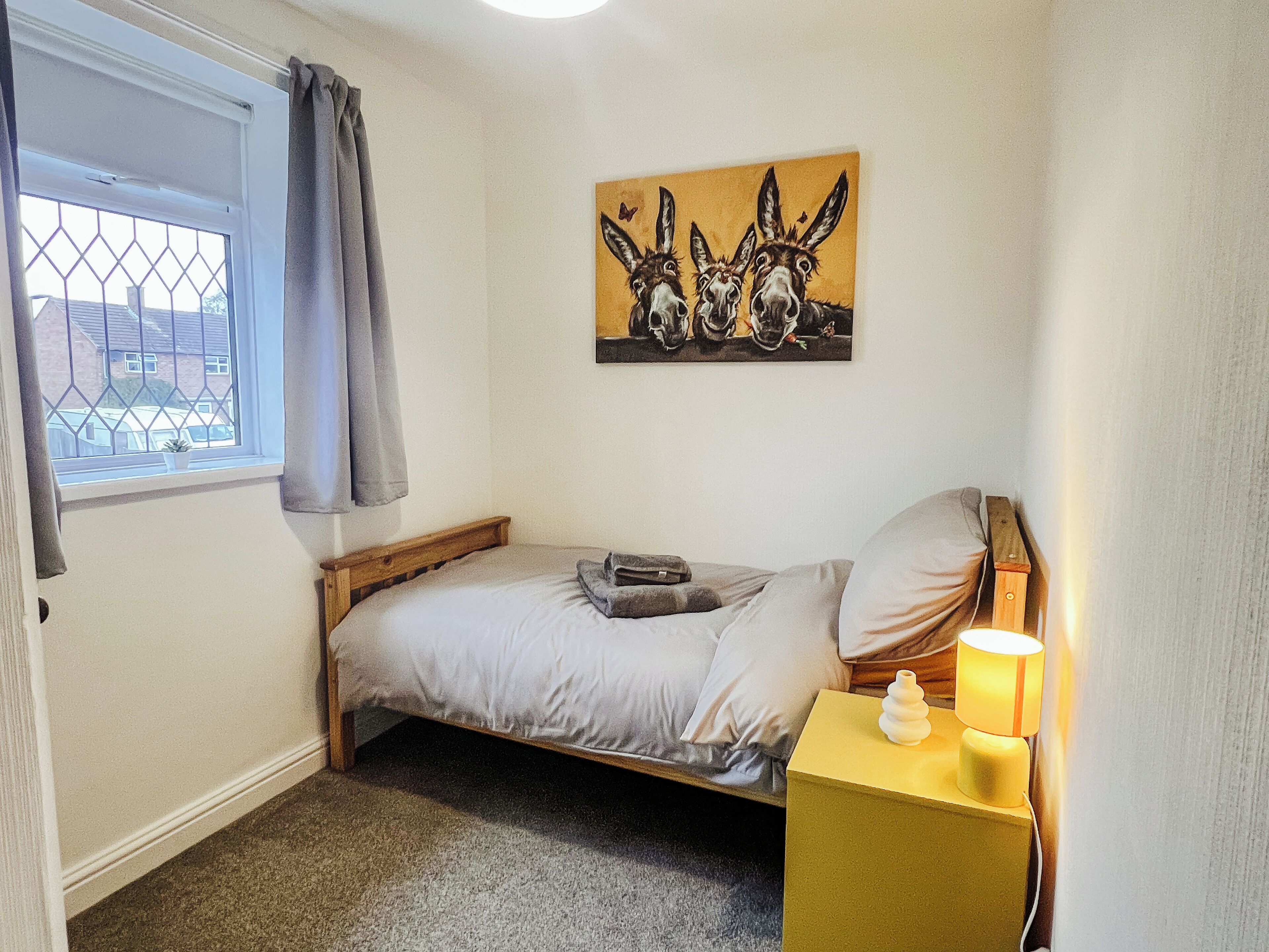 Een slaapkamer met een enkel bed, een nachtkastje met een lamp, een raam met gordijnen en een schilderij van ezels.