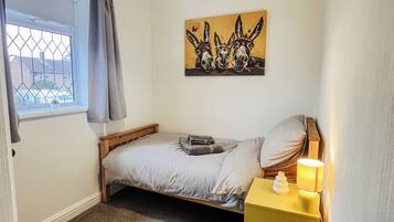 Een slaapkamer met een enkel bed, een nachtkastje met een lamp, een raam met gordijnen en een schilderij van ezels.