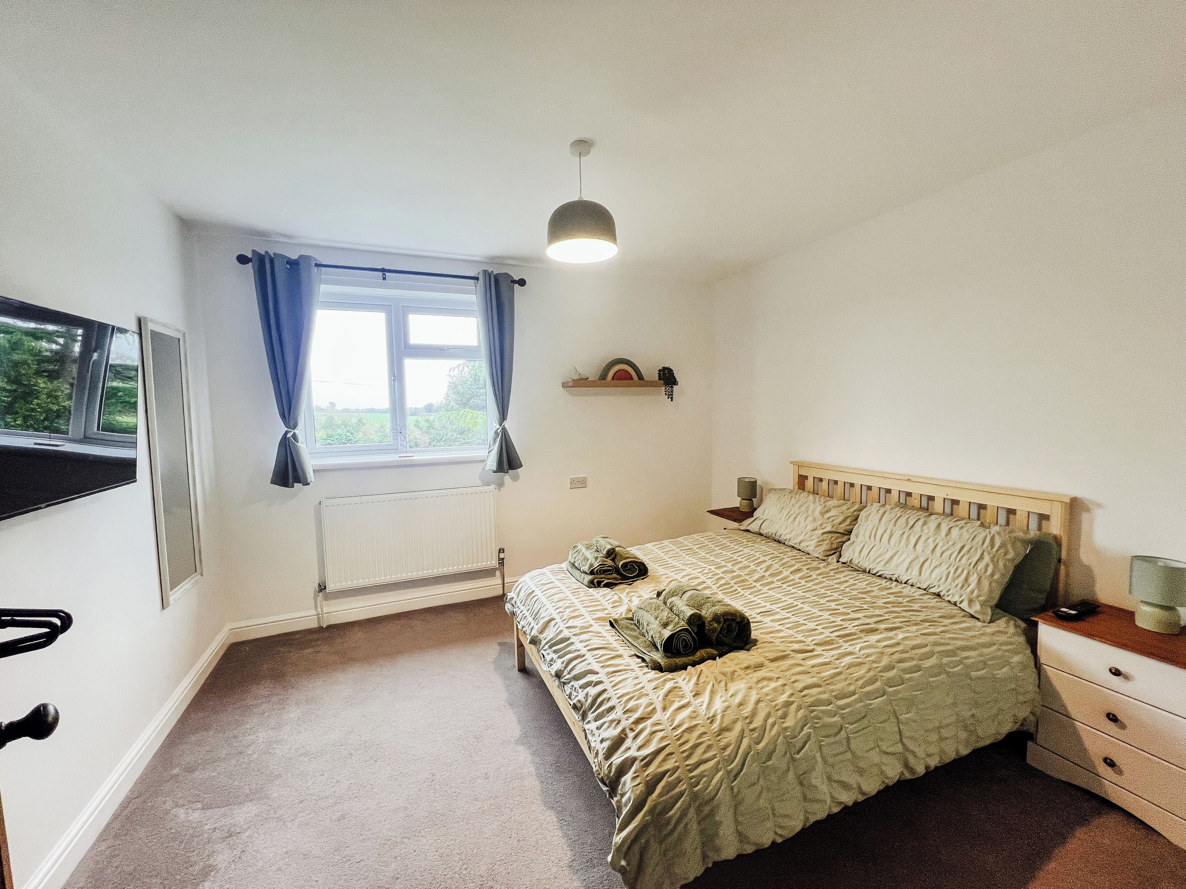 A bedroom with a bed, a window with curtains, a radiator, a bedside table with a lamp, and a shelf with decorative items.