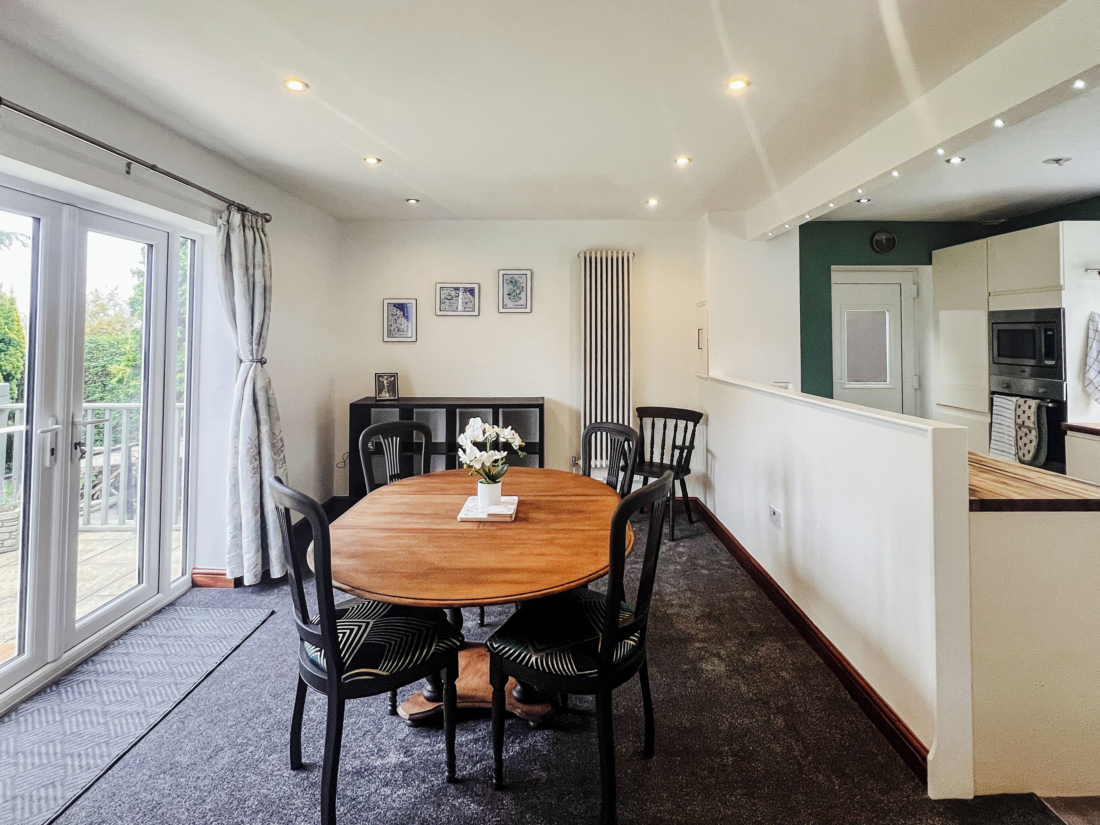 Un coin repas avec une table ronde en bois, des chaises noires et un vase rempli de fleurs. On peut voir des tableaux encadrés au mur, un radiateur et un coin cuisine à travers une porte ouverte.