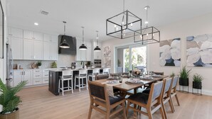 Una cocina moderna con armarios blancos, una isla central, iluminación colgante y un comedor con sillas de madera y una mesa preparada para cuatro.