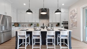 Una cocina moderna con armarios blancos, una isla central con taburetes de bar, electrodomésticos de acero inoxidable, iluminación colgante y obras de arte en las paredes.
