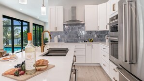 Una cocina moderna con armarios blancos, electrodomésticos de acero inoxidable y un amplio ventanal con vista a una piscina.