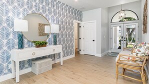 Un interior moderno con un papel tapiz estampado, una consola blanca con objetos decorativos, una silla de mimbre y una puerta que da a otra habitación.