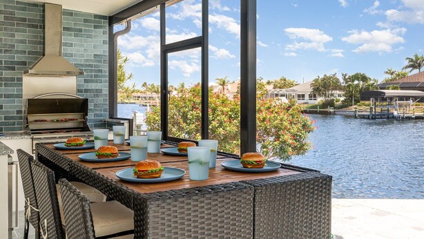 Un área de comedor al aire libre con una parrilla, sillas de mimbre y vistas a un cuerpo de agua y edificios.