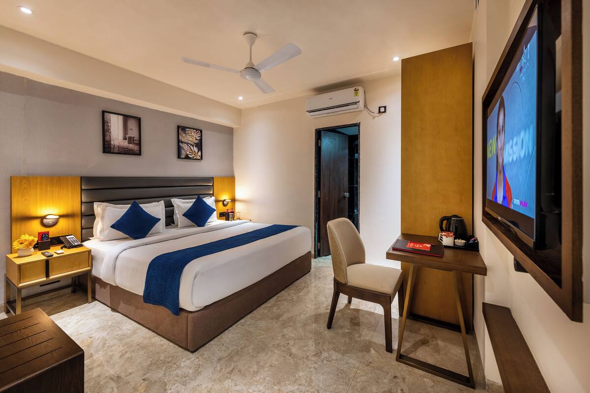A hotel room with a bed, bedside tables, a desk, a chair, a TV, and an air conditioning unit.