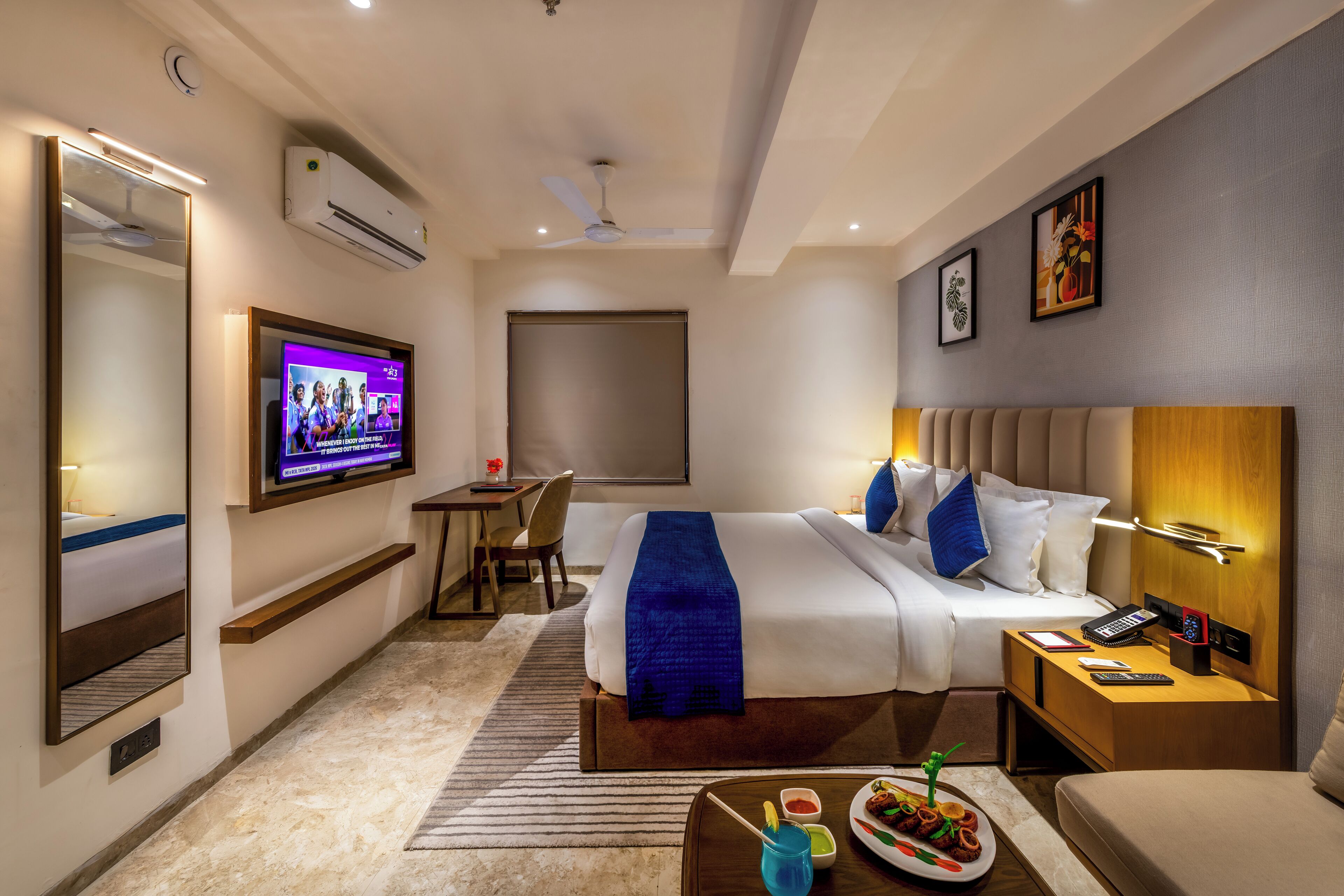 A hotel room with a bed, bedside table, TV, desk, chair, and a large mirror.