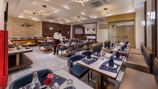 A restaurant with multiple dining tables and chairs, a chef preparing food, and a counter with various dishes. - Lords Studio Inn, Jamnagar (Jamnagar)