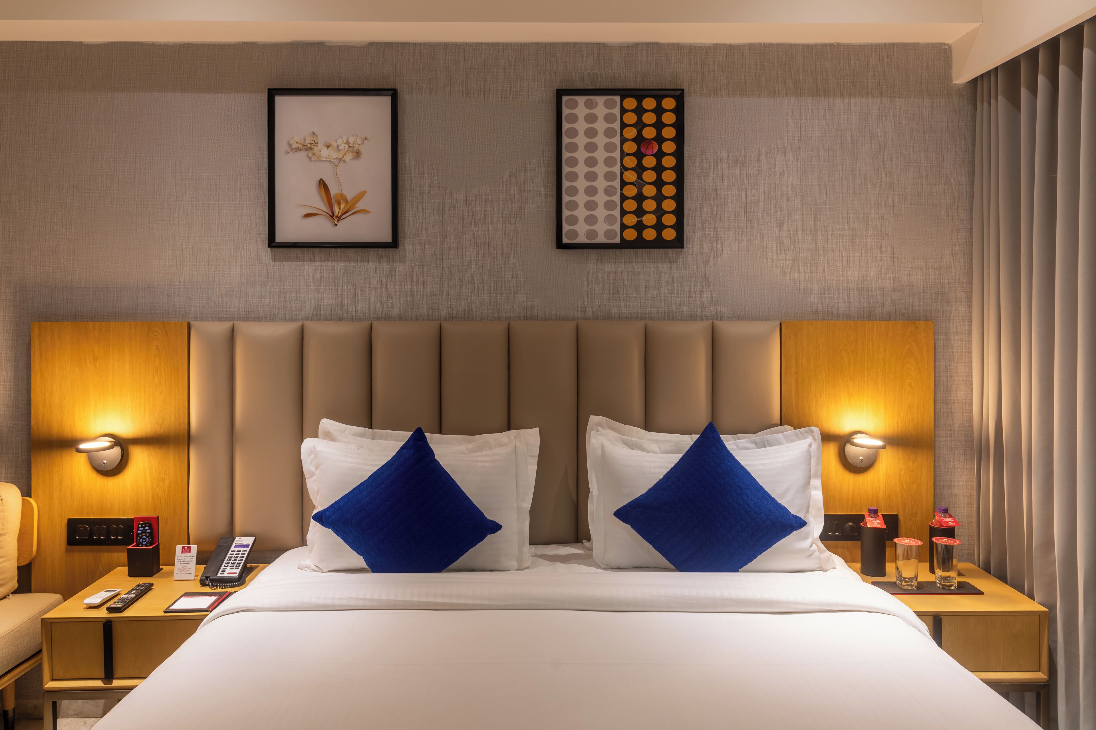 A hotel room with a large bed, two bedside tables, a headboard, wall-mounted lights, a framed picture, and a decorative wall piece.