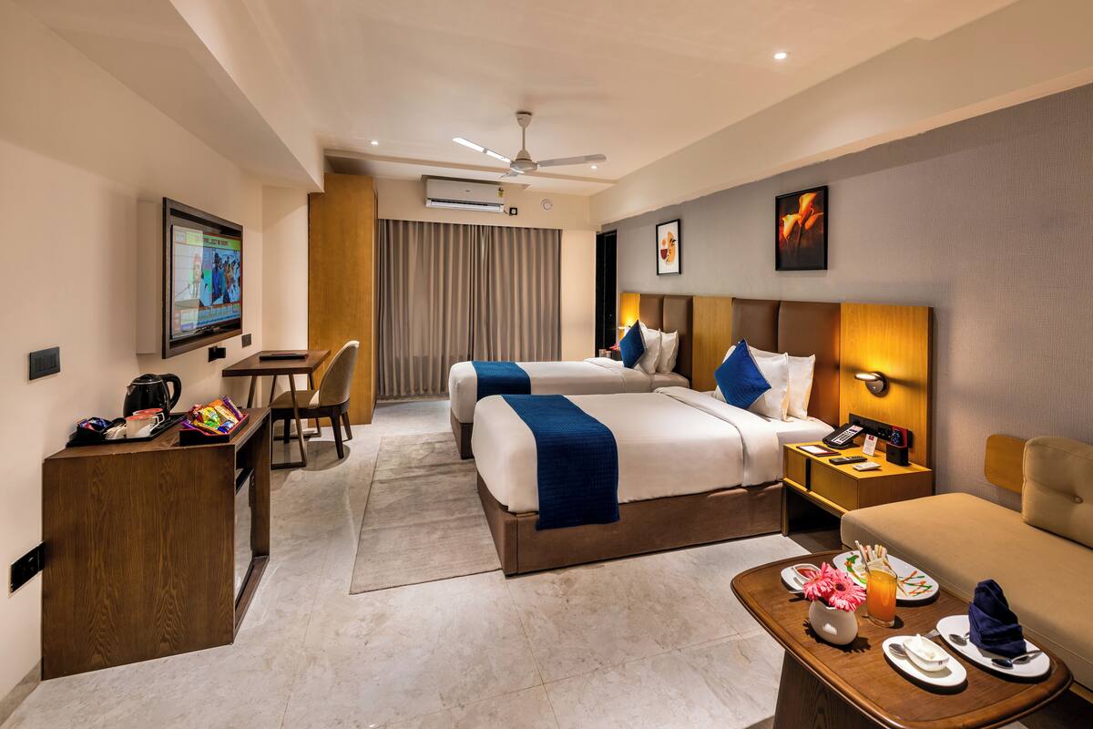 A hotel room with two beds, a desk, a chair, a coffee machine, a TV, and a dining area with plates and cutlery.