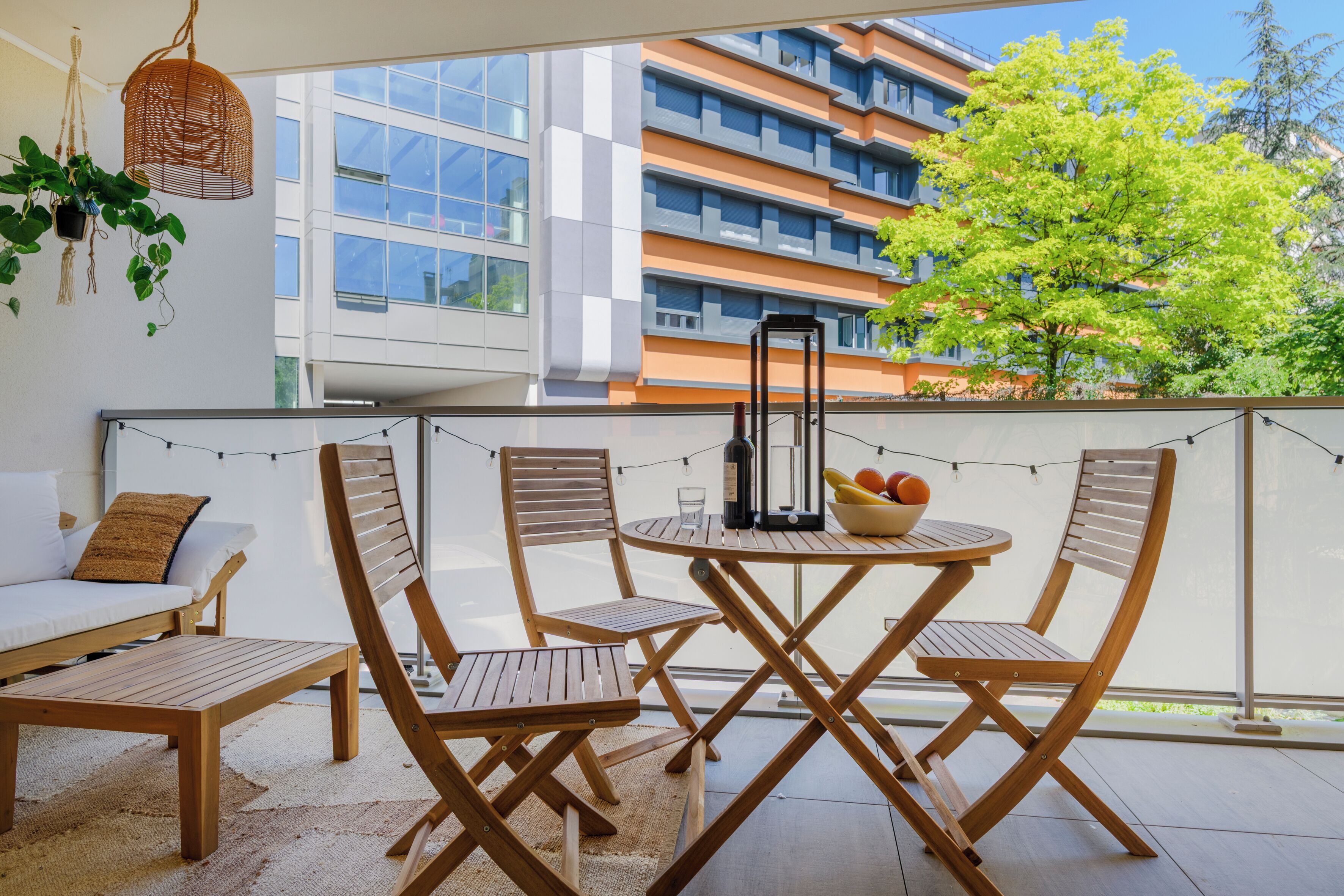 A balcony with wooden furniture, a table, and chairs. There is a hanging plant and a string of lights.