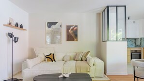 Een moderne woonkamer met een bank, een salontafel en een vloerlamp. Er is een keukenhoek met kasten en een magnetron.