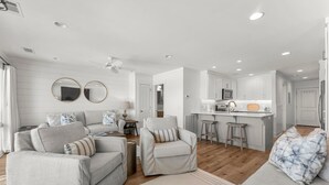 A modern living room with a sofa, armchairs, and a coffee table. There is a kitchen area with a microwave and a refrigerator.