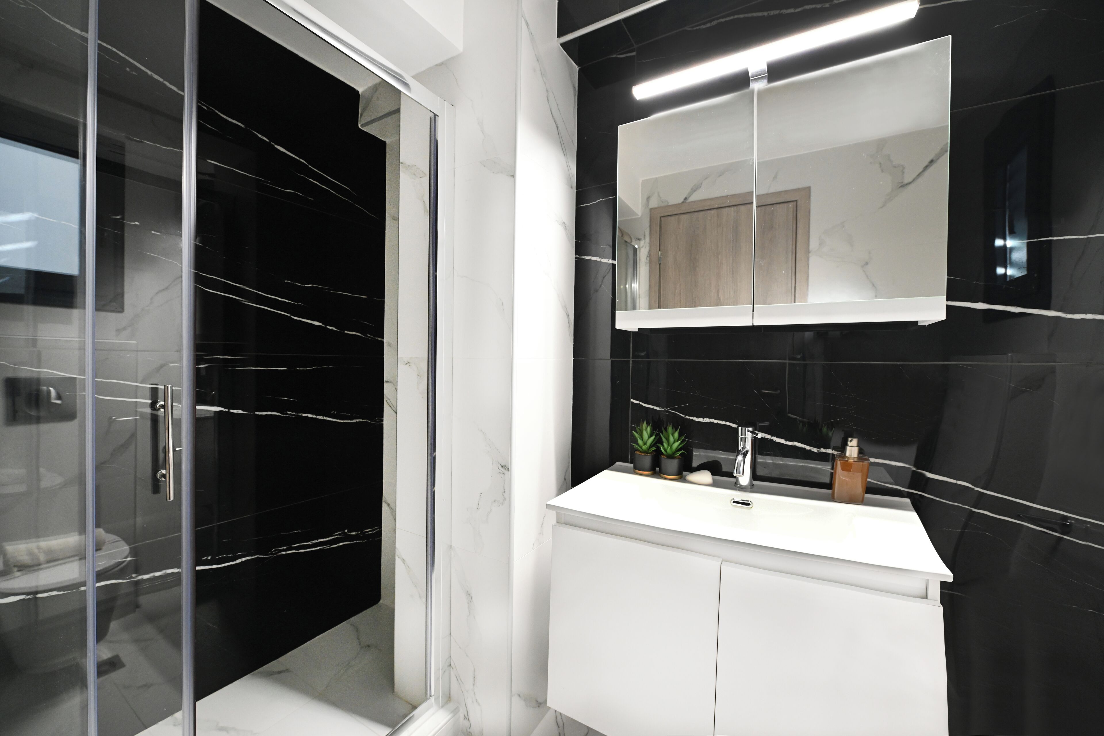 Et moderne badeværelse med en glasbrusekabine, et hvidt toiletbord med spejl og en væg med sorte fliser.