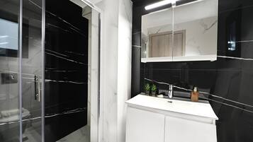 Et moderne badeværelse med en glasbrusekabine, et hvidt toiletbord med spejl og en væg med sorte fliser.