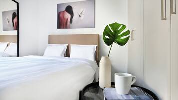 A neatly made bed with white linens, a large potted plant, a white vase, and a cup on a side table.