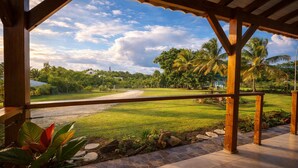 A covered outdoor area with a view of a grassy field and palm trees.