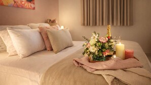 A hotel room with a bed, pillows, a floral arrangement, candles, and a bottle of champagne.