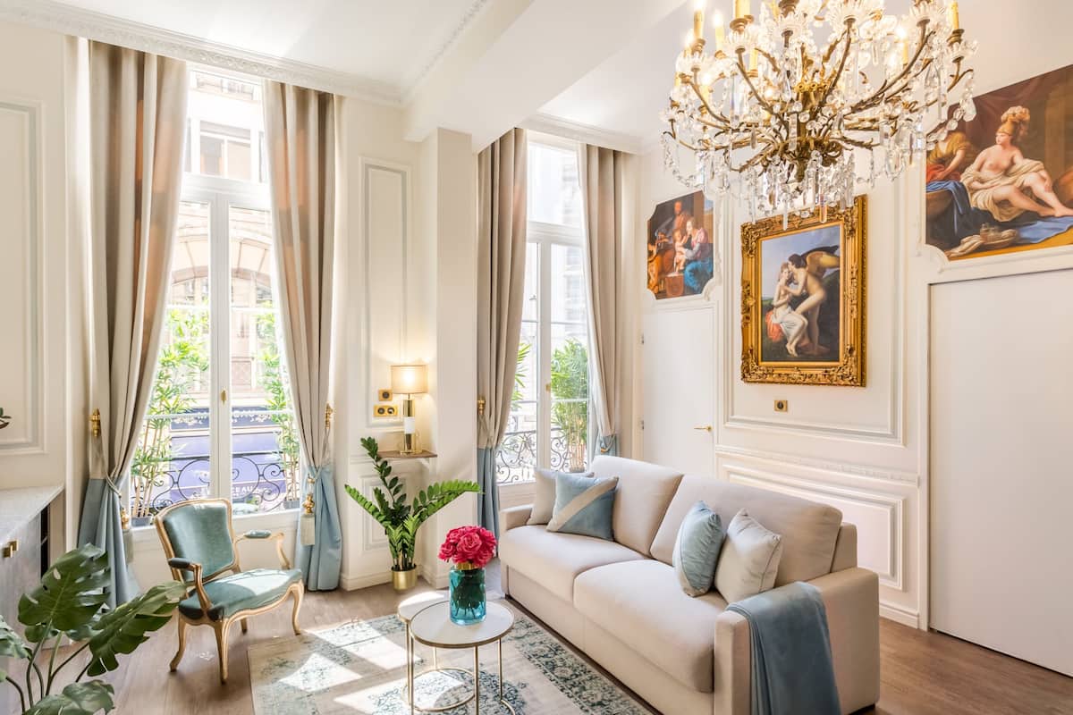 A well-lit living room with a chandelier, a sofa, a chair, and a vase of flowers.