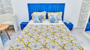 A bedroom with a blue headboard, a patterned bedspread, and matching pillows. There is a bedside table with a clock and a blue nightstand. - RESIDENCES CHELIEL, Morofé youth hostels (Yamoussoukro)