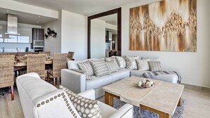 A modern living room with a sectional sofa, a dining area with wicker chairs, and a large abstract painting on the wall. - Luxe Cabo Condo w/ Ocean Views + Amenities! (Cabo San Lucas)