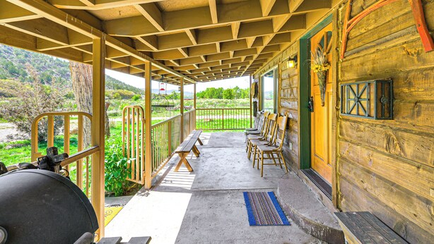A covered outdoor area with wooden benches and chairs, a grill, and a view of greenery and hills.