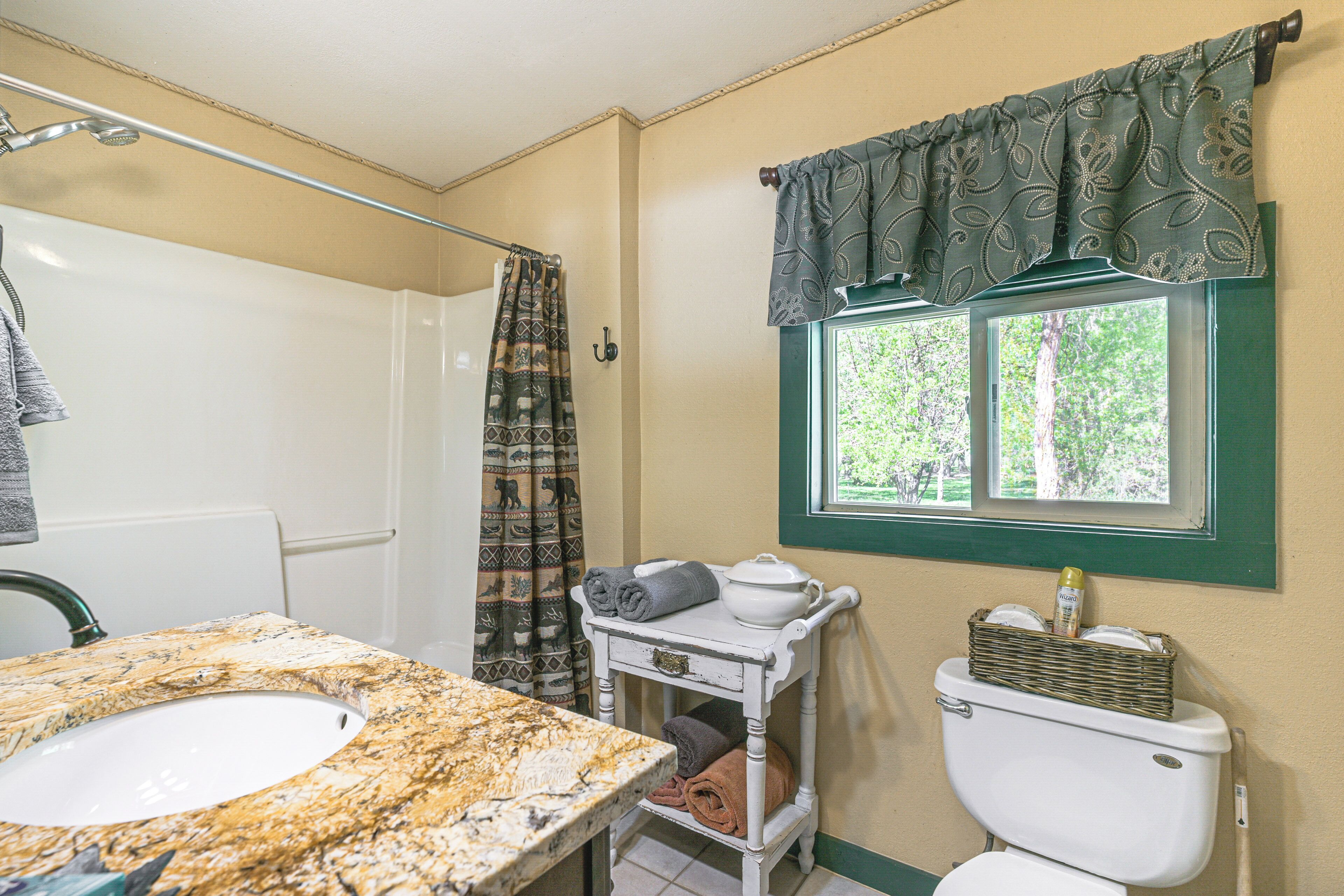 Un baño con lavamanos de mármol, inodoro blanco, una cesta de mimbre sobre el inodoro, toallero y ventana con vista a árboles.