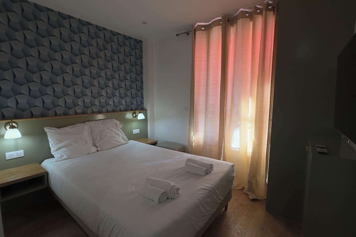 A hotel room with a bed, two bedside tables with lamps, a headboard with a geometric pattern, and a window with curtains.