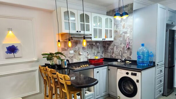 A compact kitchen with white cabinets, a built-in washing machine, and a bar area with stools. - 2 BRs Urban Oasis: Airport & Metro (Cairo Governorate)