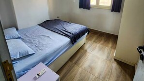 A small, neatly arranged room with a bed, a window with curtains, a ceiling light, and a small table with a remote control. - 2 BRs Urban Oasis: Airport & Metro (Cairo Governorate)