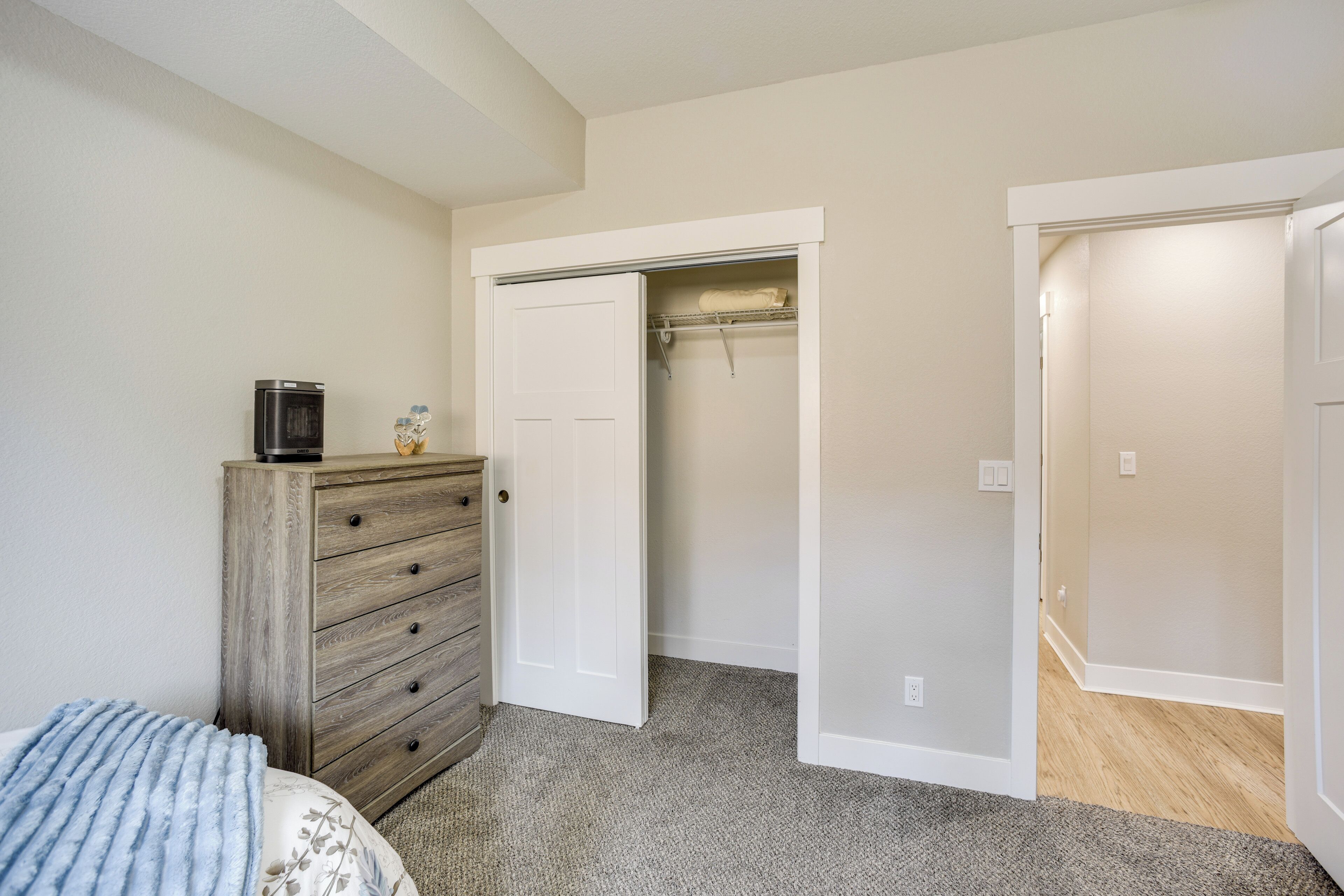 A bedroom with a wooden dresser, a bed with a floral bedspread, a closet with a hanging rod, and a door leading to another room.
