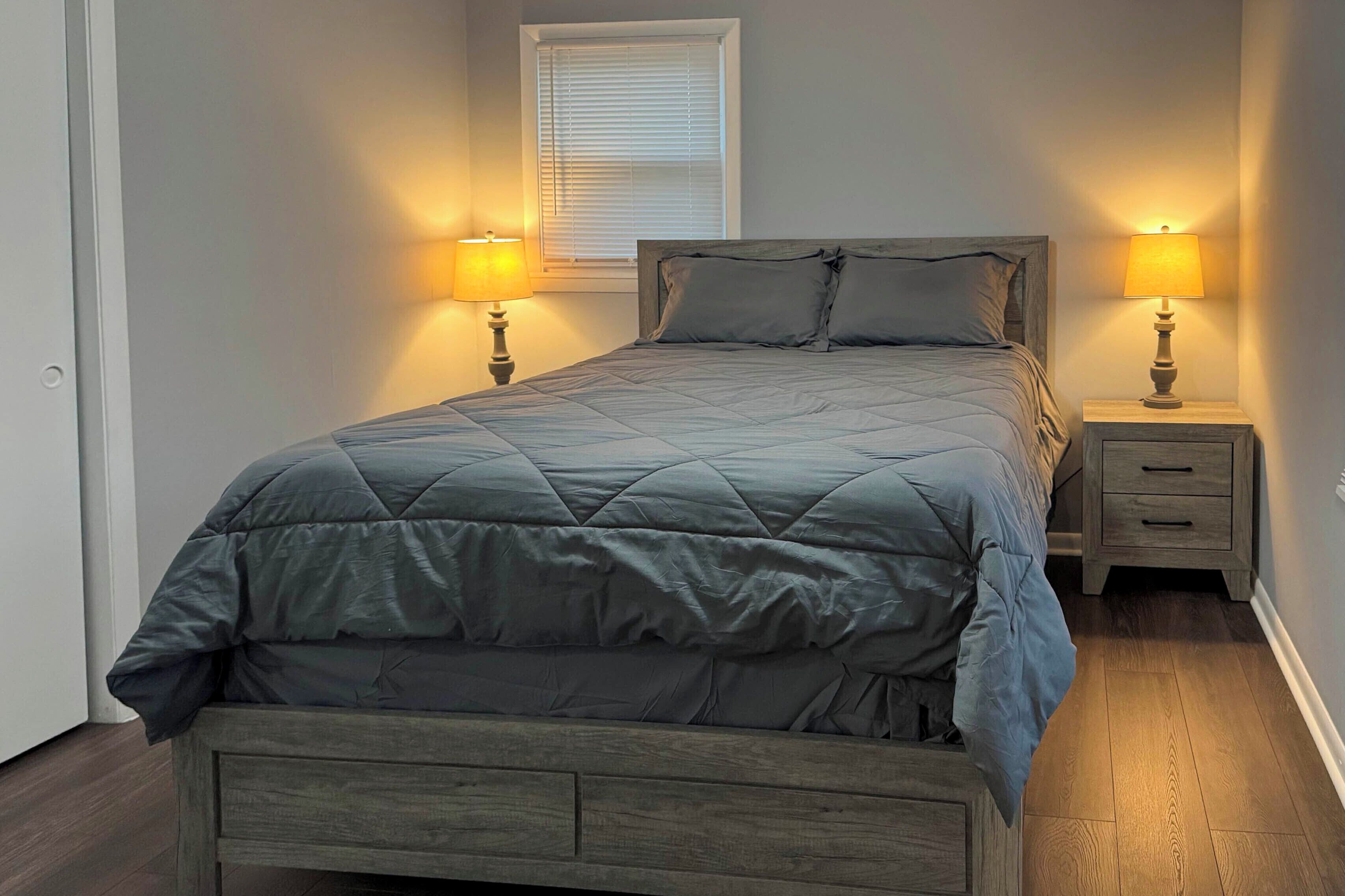 A bedroom with a bed, two bedside tables with lamps, a window with blinds, and a door.