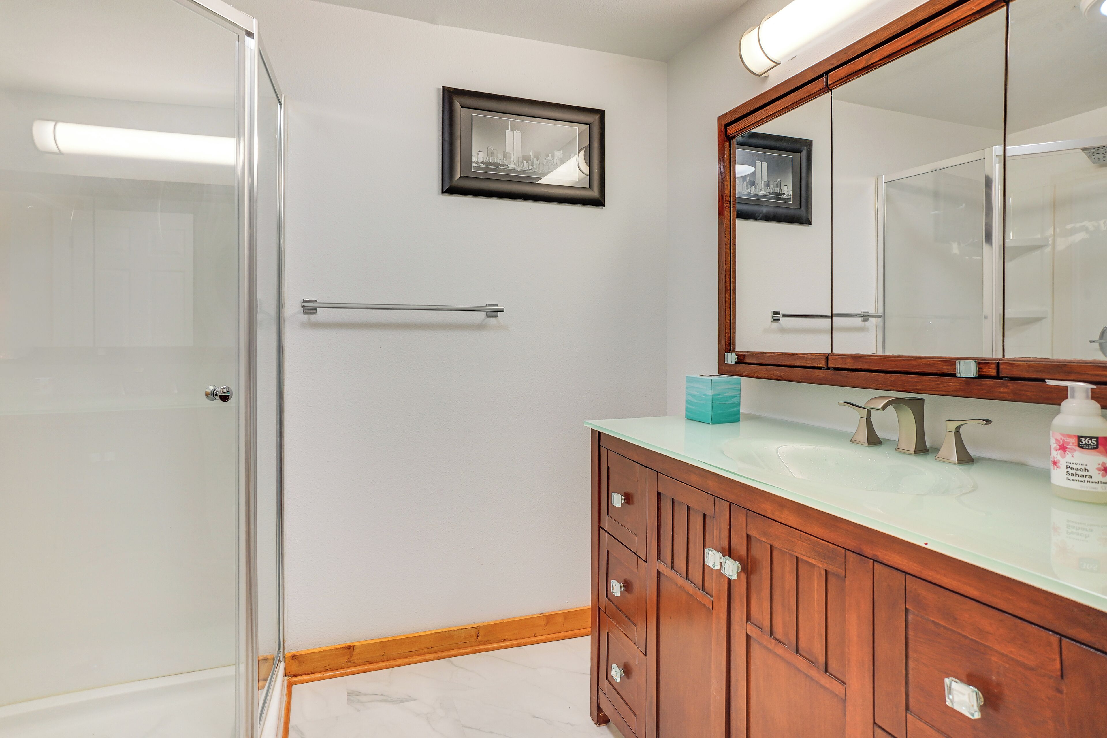 A bathroom with a glass shower enclosure, wooden vanity with a mirror, and a framed picture on the wall.