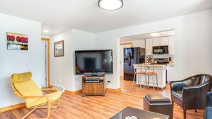 A modern living room with a flat-screen TV, yellow and black leather chairs, a coffee table, and a kitchen area with white cabinets and a microwave. - 1 Mi to Shore: Lake Michigan Retreat w/ Game Room! (Two Rivers)