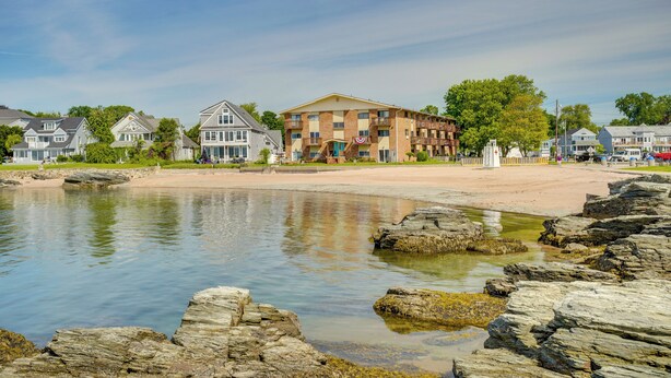 Un hôtel situé sur la côte, avec une plage de sable, une côte rocheuse et, en arrière-plan, un mélange de bâtiments résidentiels et commerciaux.