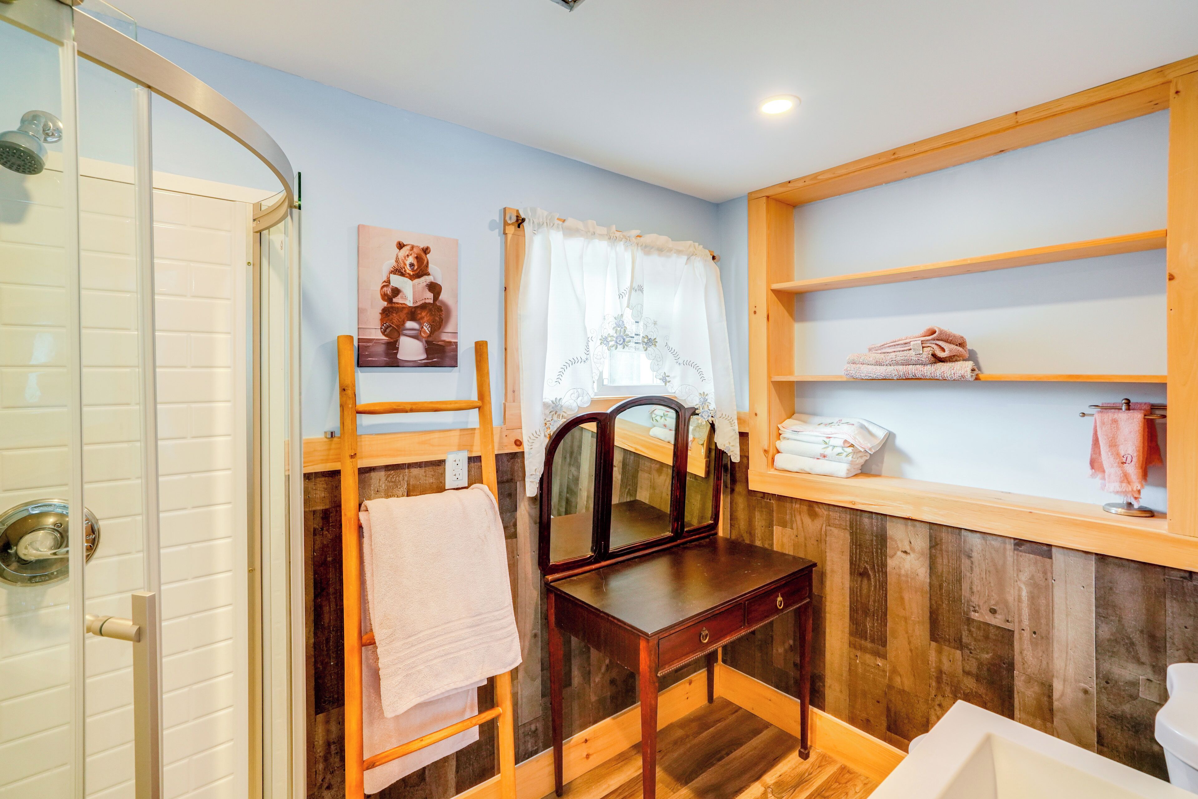 A bathroom with a wooden vanity, a towel rack, a shower with a glass door, and a shelf with towels.