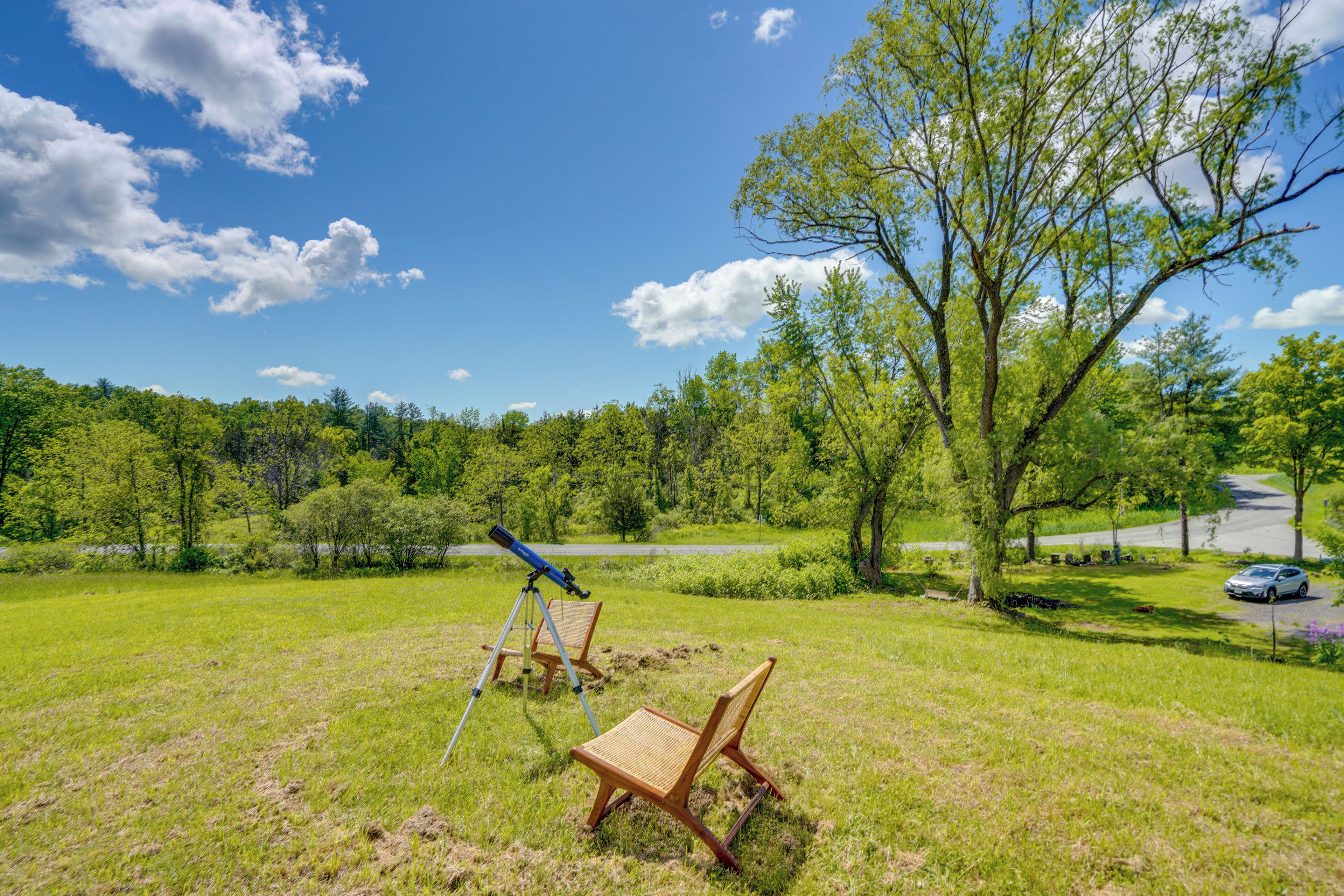 A grassy field with a telescope on a tripod and a chair, a tree, and a road with a car in the distance.