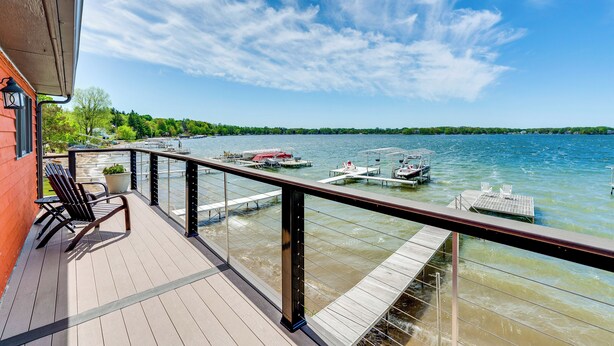 Una terraza de madera con sillas y una planta en maceta, con vistas a un lago con barcos y muelles.