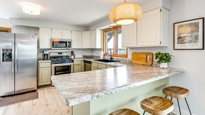 Una cocina moderna con armarios blancos, encimera de granito, refrigerador de acero inoxidable, horno empotrado y lámpara colgante.
