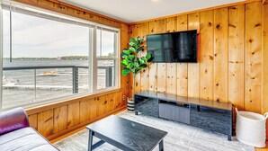 Una habitación con un ventanal grande con vista a un cuerpo de agua, un televisor de pantalla plana en la pared, un banco de madera y una pequeña mesa.