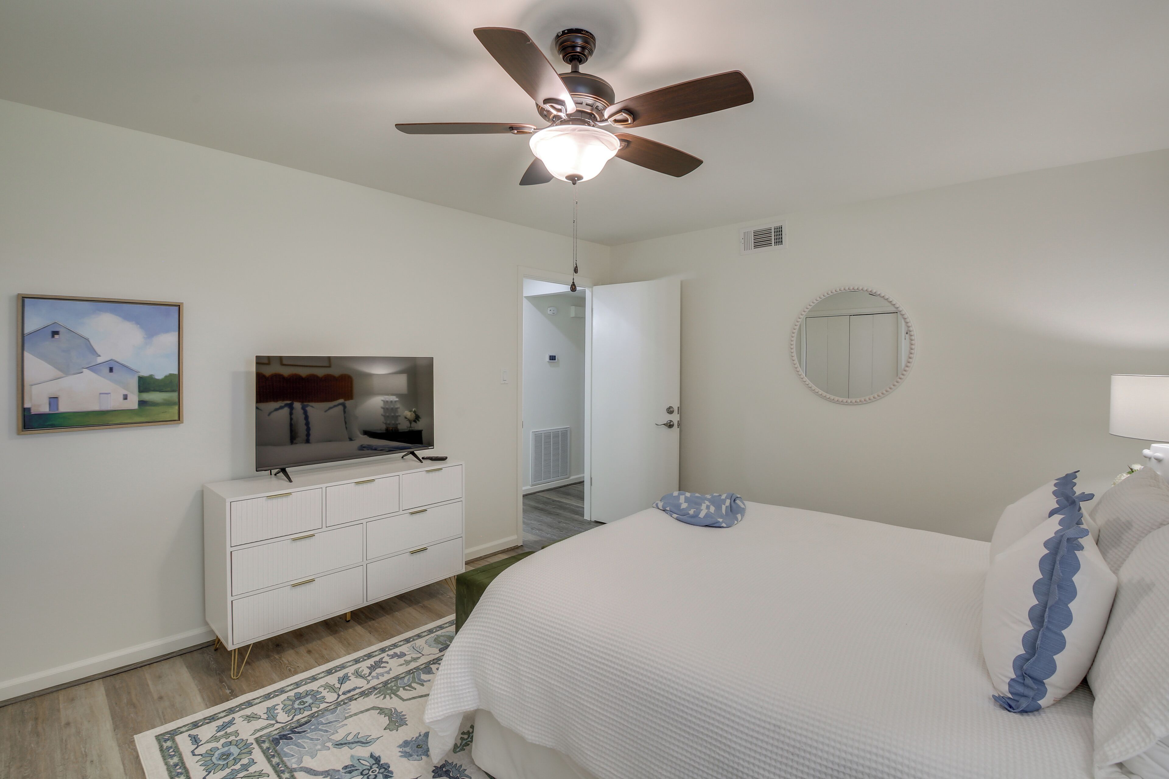 A bedroom with a bed, a dresser, a ceiling fan, a mirror, and a painting on the wall.